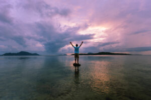 Man standing on stone in water-purple sunrise
